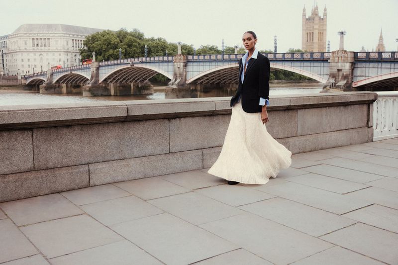 Model standing on a stone embankment wearing a black blazer, blue shirt, and long white skirt with a bridge in background.