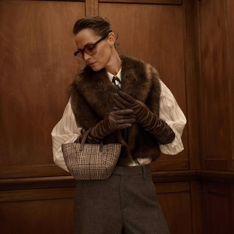 Model wearing fur vest and leather gloves holding a plaid mini bag inside a wood-paneled elevator interior.