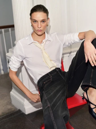 Model seated on a red chair wearing a white shirt, cream tie, and black trousers against a neutral interior background
