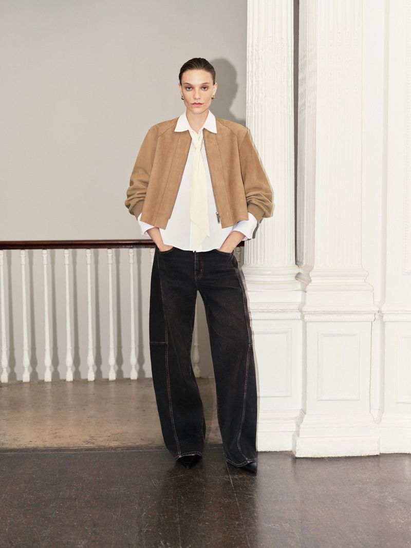 Model wearing a tan jacket, white shirt, and black wide-leg trousers standing indoors next to a white pillar.