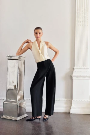 Model wearing cream halter top and black wide-leg trousers leaning against a metallic pedestal in an interior space