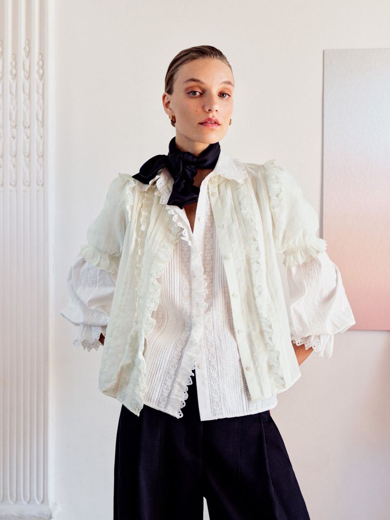Model in white ruffled blouse and black trousers wearing a black silk neck scarf against a white interior wall.