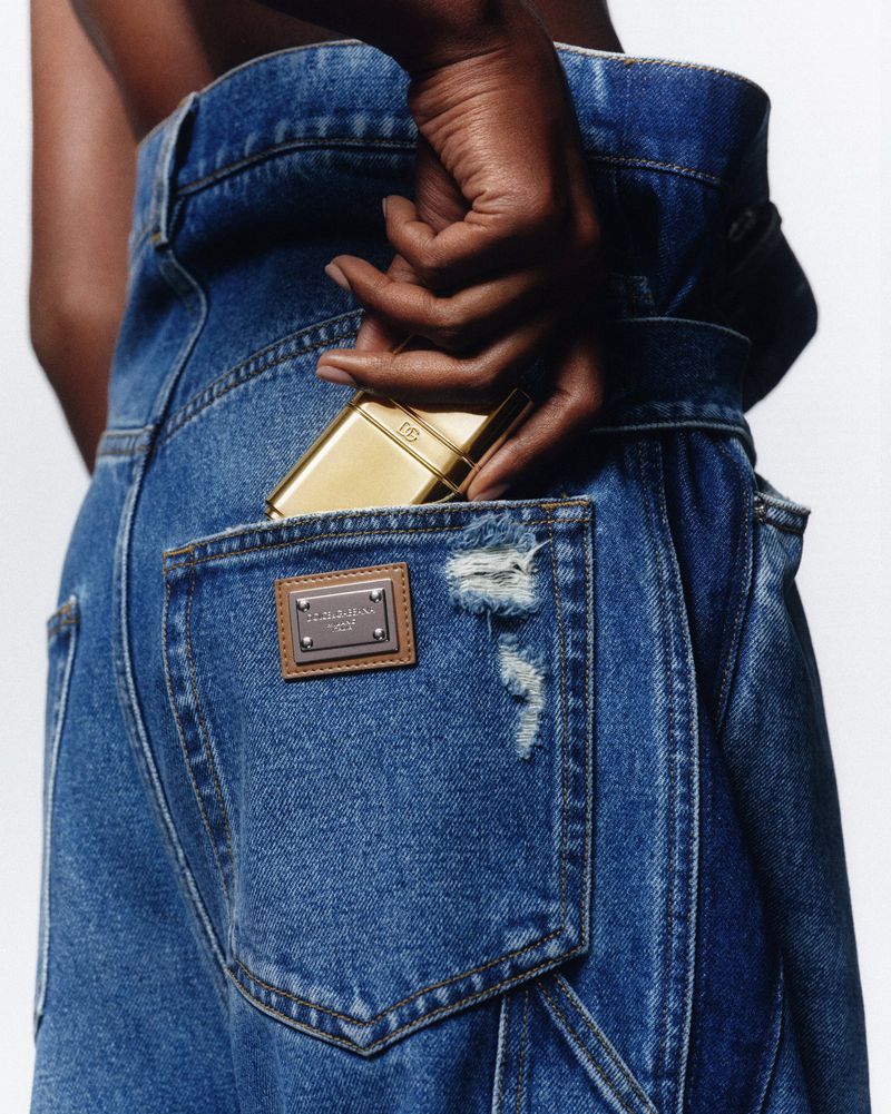 Close-up of a person sliding a gold compact into the back pocket of distressed denim jeans against a white background