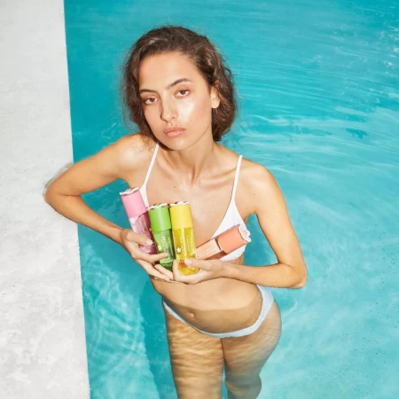 Model in white swimwear standing in turquoise swimming pool holding colourful Marc Jacobs Beauty perfume bottles