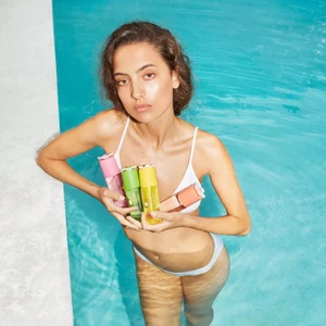 Model in white swimwear standing in turquoise swimming pool holding colourful Marc Jacobs Beauty perfume bottles