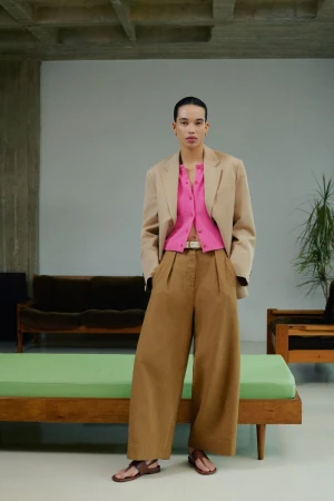 Model standing wearing a beige blazer, pink button-down shirt, and brown wide-leg trousers in a modern interior.