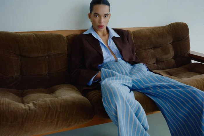 Model seated on a brown corduroy sofa wearing a blue striped trousers, light blue shirt, and dark brown blazer.