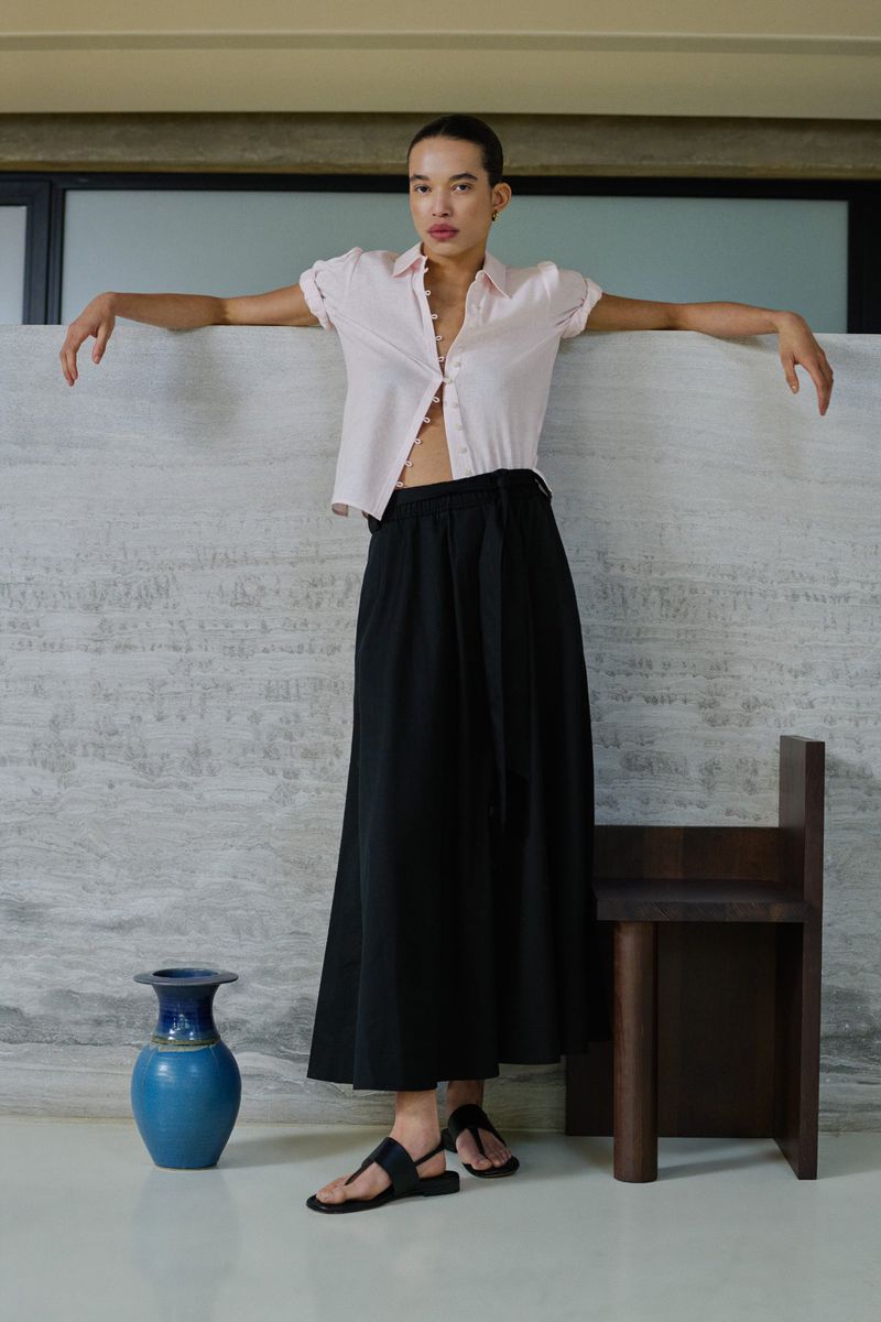 Model Lara Menezes wearing a pink button-down shirt and black skirt leans against a textured stone wall in a ME+EM lookbook look.