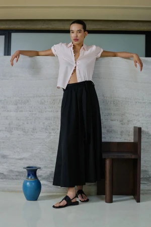 Model Lara Menezes wearing a pink button-down shirt and black skirt leans against a textured stone wall in a ME+EM lookbook look.