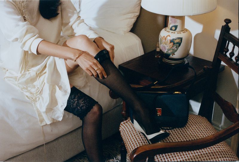 Model seated on a bed pulling up black sheer stockings, wearing an off-white dress in a hotel room setting.