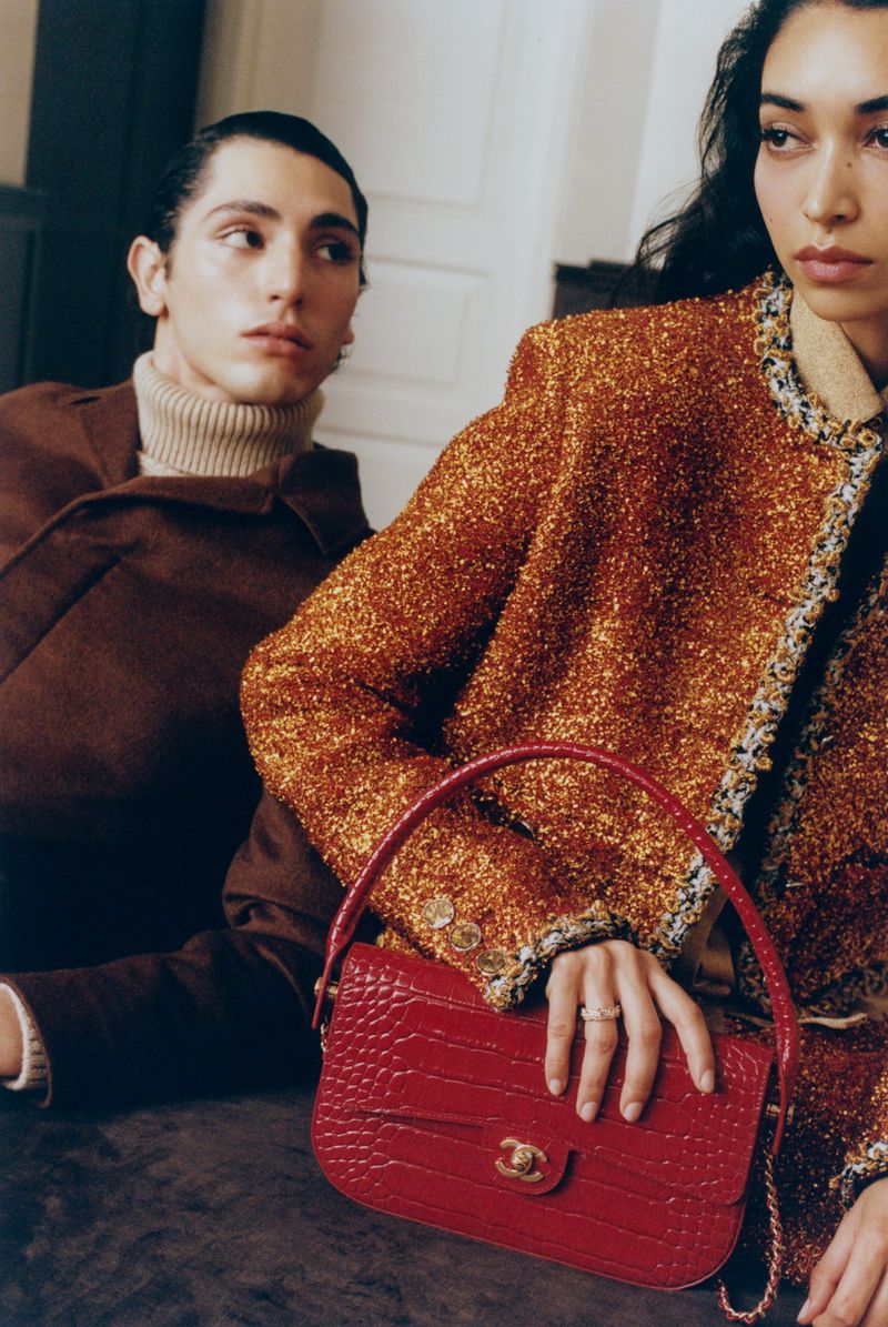 Two models posing together, woman in shimmering gold tweed jacket resting hand on red leather handbag, man in brown coat and turtleneck