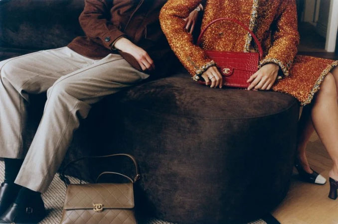 Two seated models wearing a brown jacket and a textured orange coat holding a red handbag for Vogue Latin America.