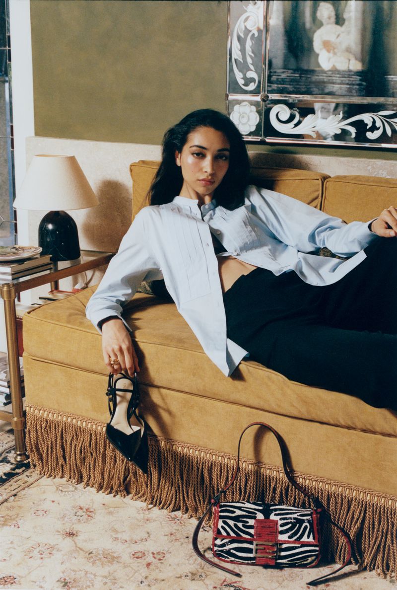 Model reclining on a mustard yellow velvet sofa holding a black high-heeled shoe, with a zebra-print handbag on the floor.
