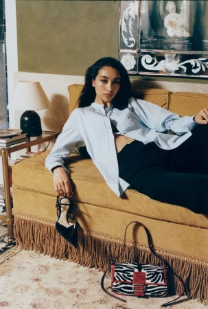 Model reclining on a mustard yellow velvet sofa holding a black high-heeled shoe, with a zebra-print handbag on the floor.