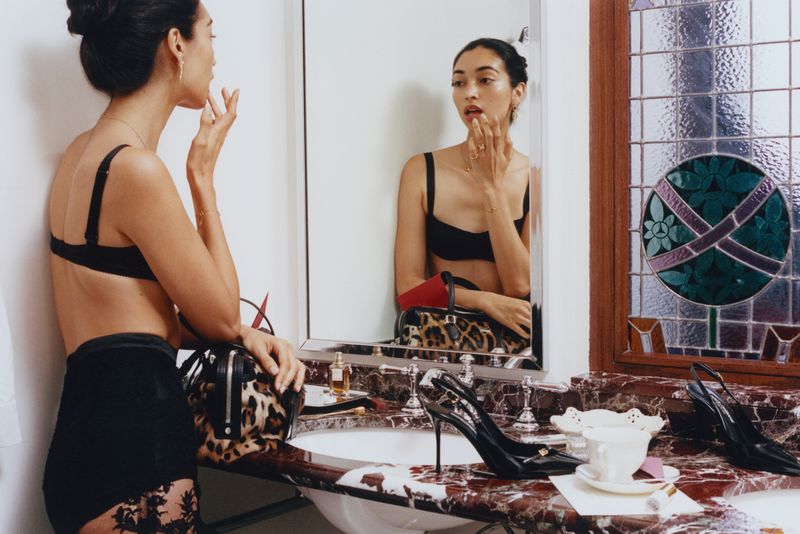 Model in black lingerie standing before a bathroom mirror applying lipstick for a Vogue Latin America editorial.