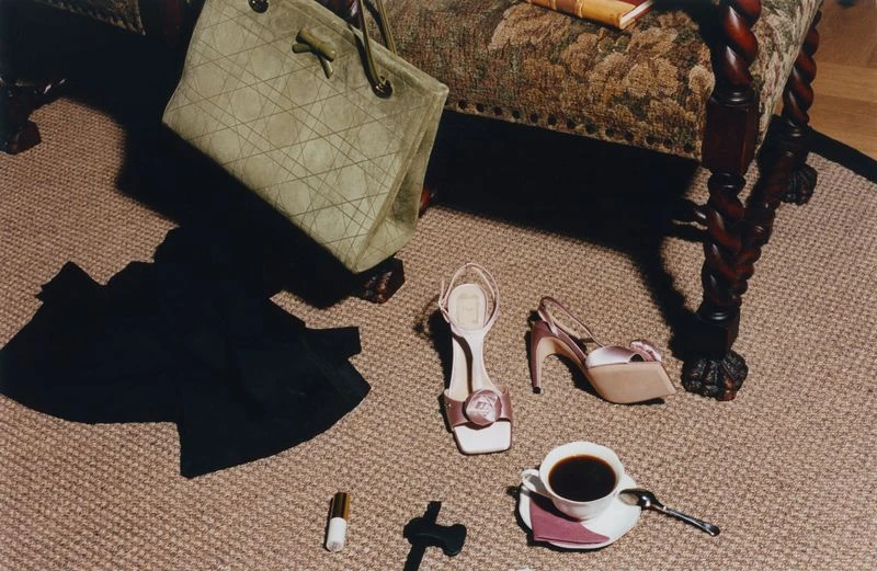 Olive green quilted tote bag, pink high-heeled sandals, dark clothing, and coffee cup scattered on a textured carpet.