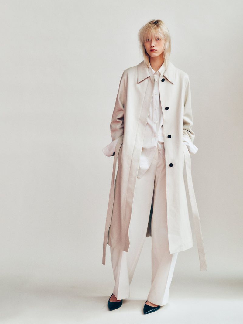 Model wearing a long beige trench coat over matching trousers against a pale seamless studio background.