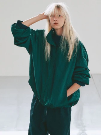 Model with long blonde hair wearing a green windbreaker jacket standing against a white seamless background.