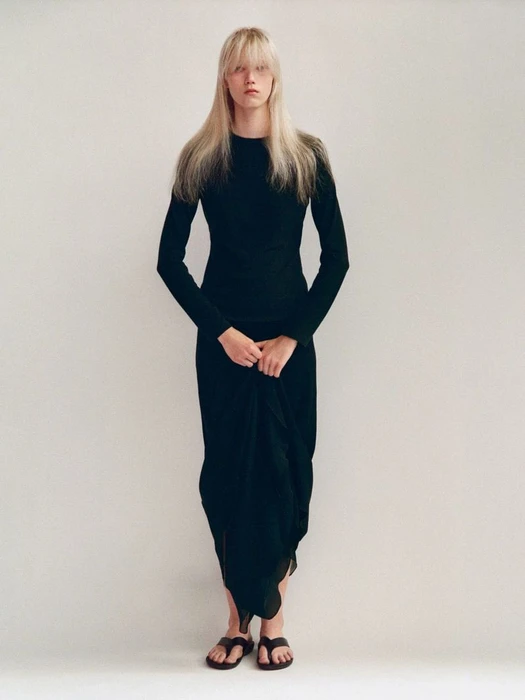 Model with long blonde hair wearing a long black dress and dark sandals against a neutral studio background