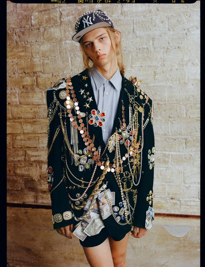 A young male model wearing an embellished dark blazer covered in chains and jewels and a baseball cap against a brick wall background