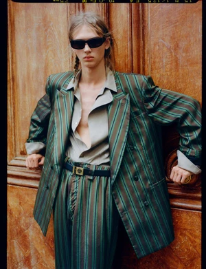 Model wearing a striped green blazer and matching trousers leaning against a dark wood door in HERO Magazine