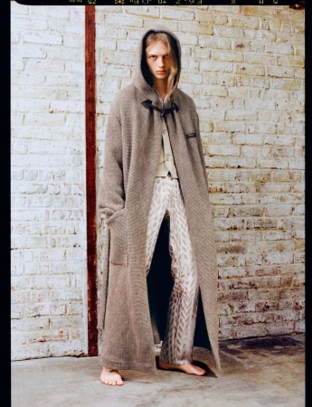 Model in a long grey hooded coat standing against an exposed white brick wall.