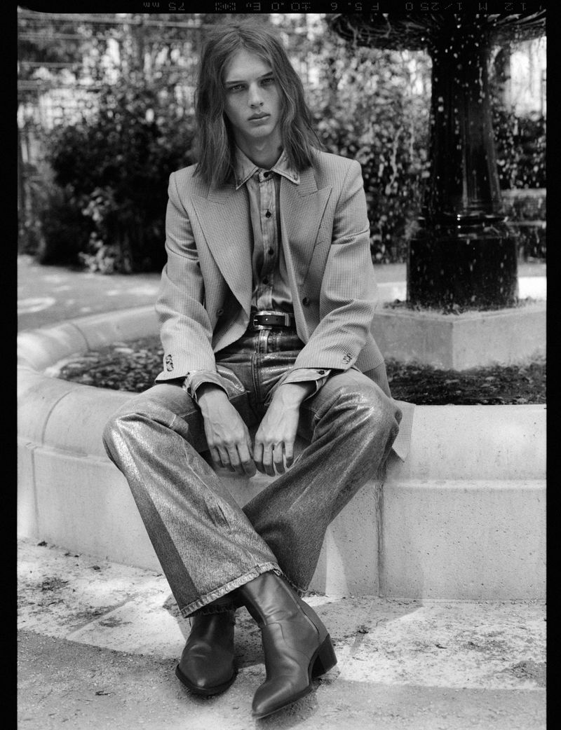 Young male model with long hair wearing a tailored blazer and boots seated outdoors near a stone fountain