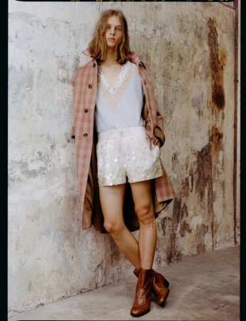 Model in plaid trench coat, light V-neck top, patterned shorts, and brown boots leaning against a distressed wall.