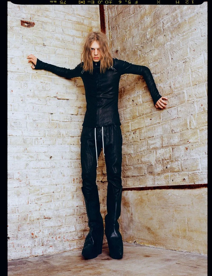 A long-haired male model in a black leather jacket and boots standing against an exposed brick wall corner.