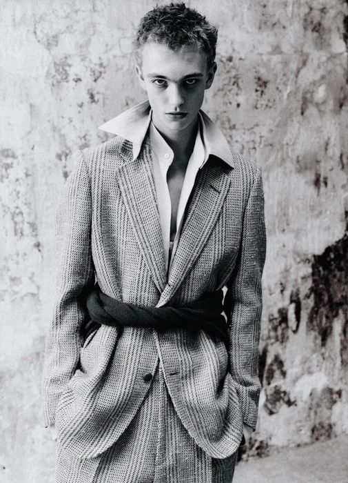 Young male model wearing a textured patterned blazer and white shirt against a weathered textured wall