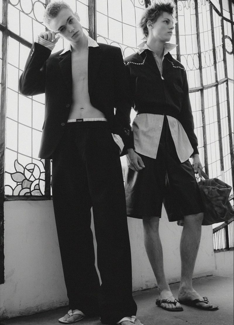 Two young male models in black jackets and trousers standing against large decorative glass windows in black and white