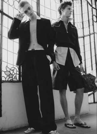 Two young male models in black jackets and trousers standing against large decorative glass windows in black and white