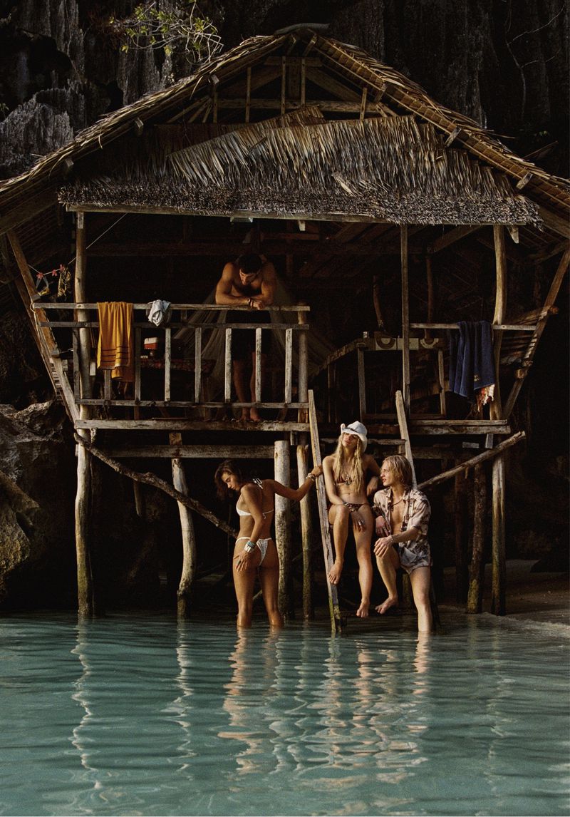 Several people relax by a rustic stilt hut on a tropical beach with clear turquoise water and dark, rocky cliffs in the background.