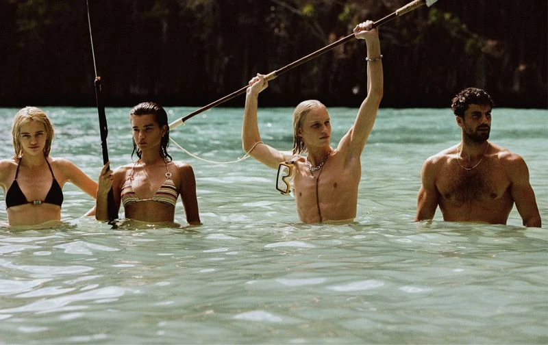 Four people wade waist-deep in the turquoise water of a tropical beach. Two women in bikinis flank two shirtless men near the dark shoreline.