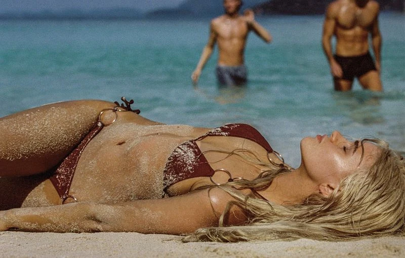 Woman in bikini lying on sandy beach with two men standing in turquoise water in background under natural light during the daytime.