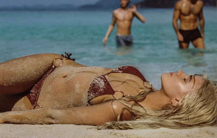Woman in bikini lying on sandy beach with two men standing in turquoise water in background under natural light during the daytime.