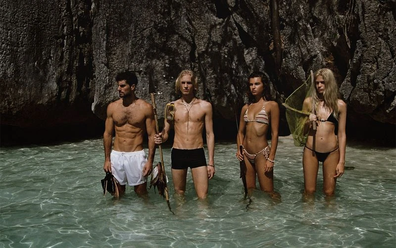 Four attractive young adults stand waist-deep in clear water near a rocky shore. Two men, two women wearing swimwear. Spear fishing theme.