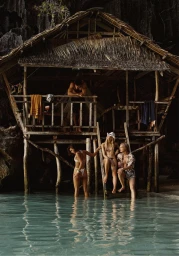 Four models by a thatched stilt hut over shallow turquoise water with dramatic rock cliffs behind.