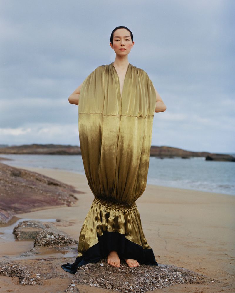 Model FeiFei Sun wearing a voluminous golden gown standing barefoot on a rocky shore for Vogue China