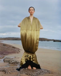 Model FeiFei Sun wearing a voluminous golden gown standing barefoot on a rocky shore for Vogue China