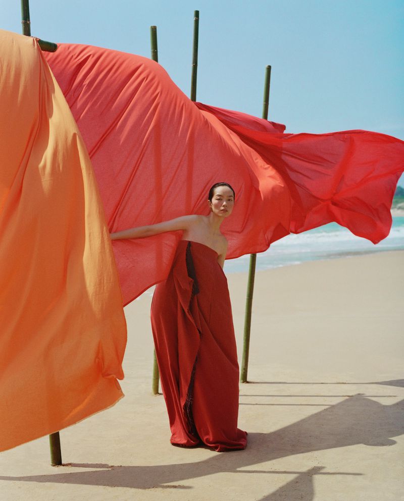 Model FeiFei Sun wearing a strapless red dress on a sandy beach next to large hanging orange and red fabric banners.
