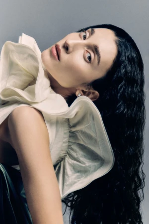 Portrait of a woman with dark wavy hair wearing a white ruffled top and green skirt against a grey background