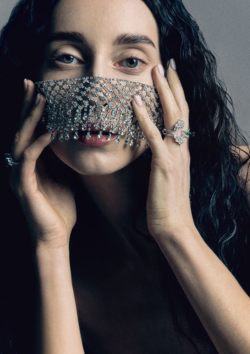 Close-up portrait of a woman wearing an intricate crystal mesh face veil, holding her hands up to her face against a neutral grey studio background