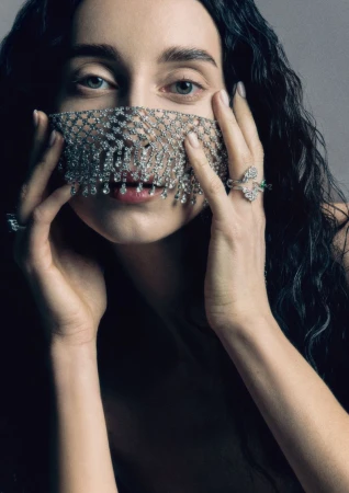 Close-up portrait of a woman wearing an intricate crystal mesh face veil, holding her hands up to her face against a neutral grey studio background