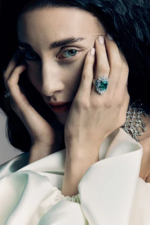 Close-up portrait of a woman wearing an emerald ring and diamond necklace with her hands framing her face.