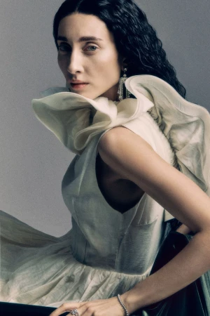 A woman with dark curly hair wearing an off-white ruffled dress against a grey studio backdrop.