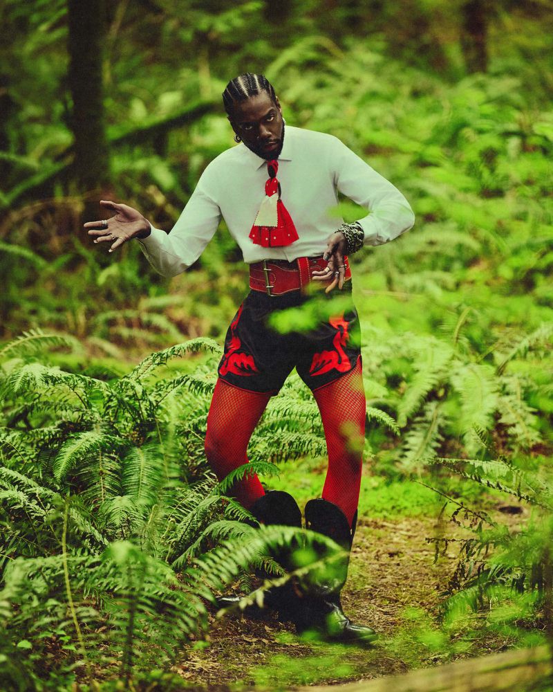 Serpentwithfeet performing in a lush green forest wearing a white shirt, red tie, black shorts, red tights, and black boots.