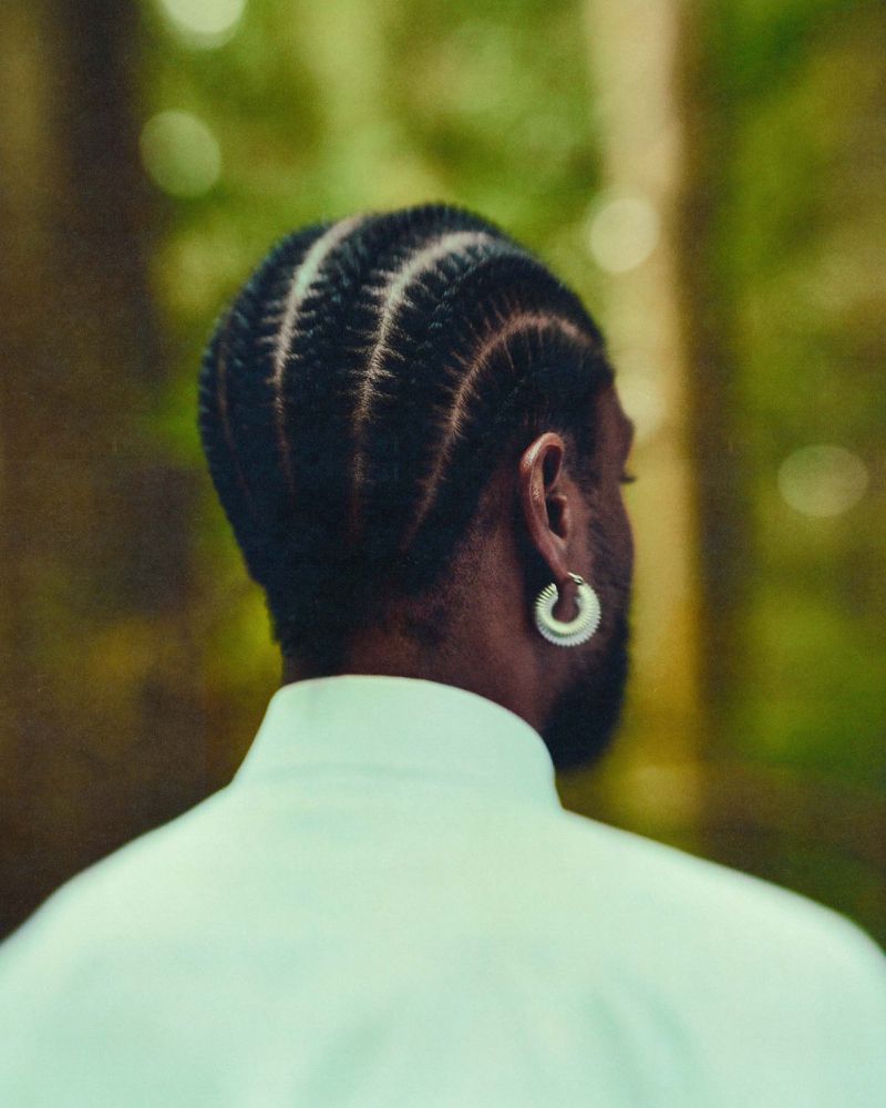 Rear view of a man with cornrows wearing a light green shirt and silver hoop earring against a blurred forest background.