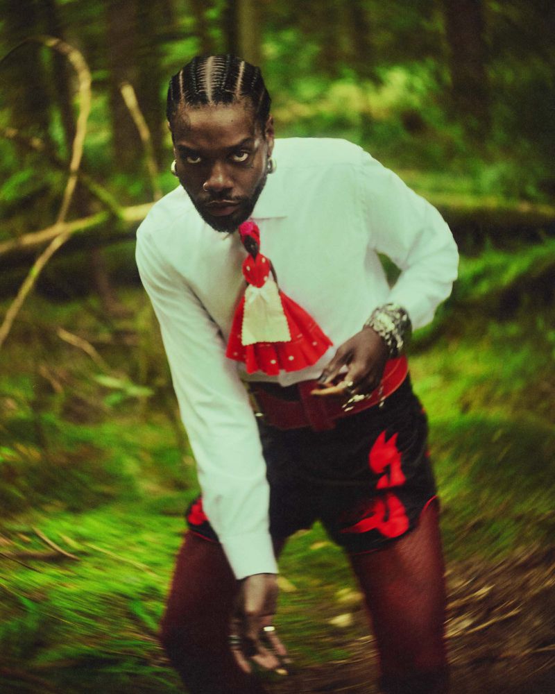 Serpentwithfeet wearing a white button-down shirt and red figurine accessory in a lush green forest