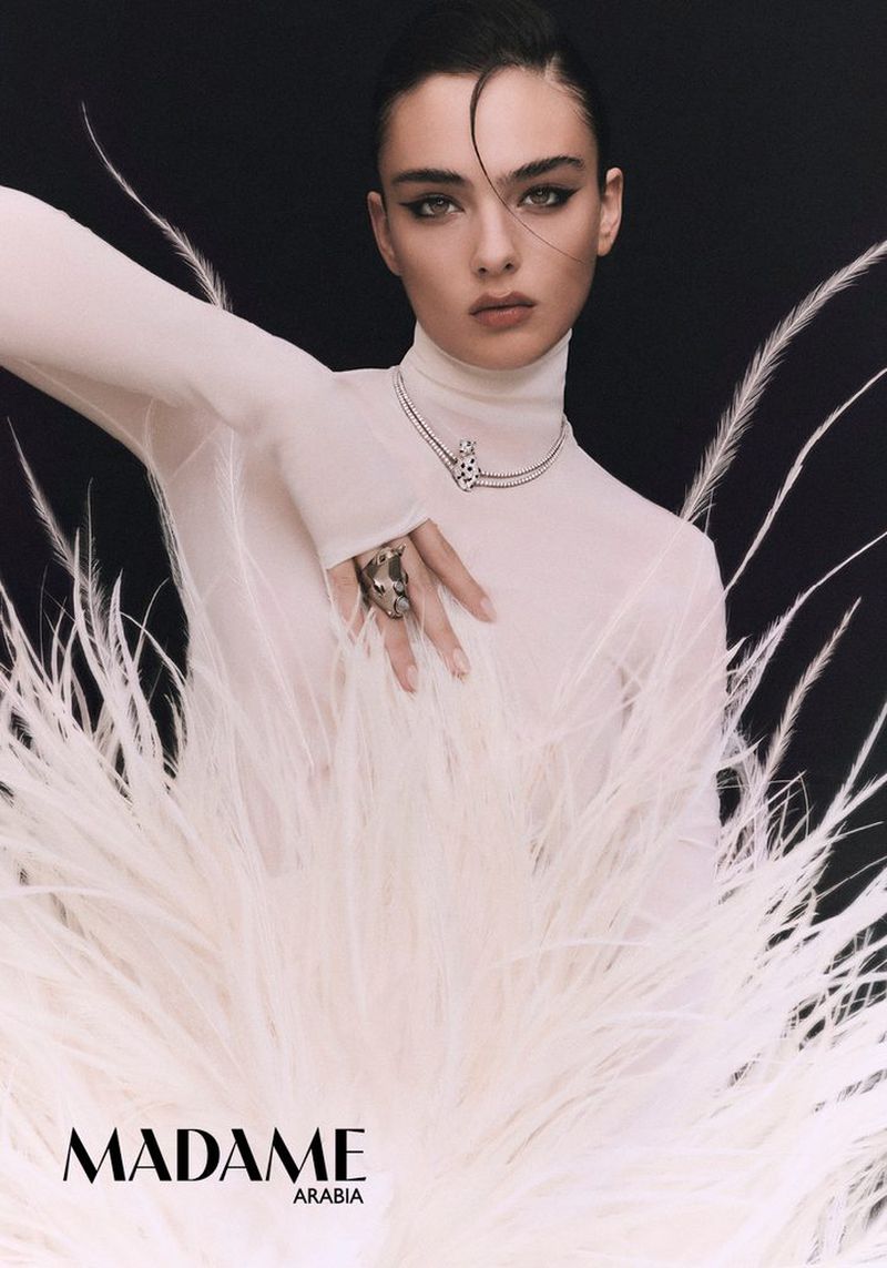 Deva Cassel in a white sheer turtleneck behind large white feathers wearing a silver ring and necklace against a dark background.